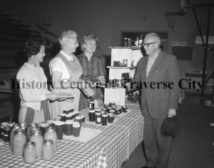 Gretchen (Arntz) Votruba, in the apron, at a St. Francis Bazaar selling canned goods. Gretechen was active in the Salvation Army and Child and Family Services and later at the Dennos Museum, ca. 1940s. Image courtesy of the History Center of Traverse City.
