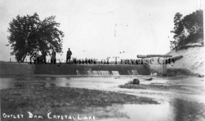 Crystal Lake outlet dam with three people standing in the lake (ca. 1920). Image courtesy of History Center of Traverse City.