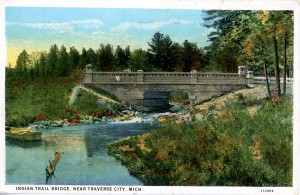 This colored postcard image depicts Indian Trail Bridge, which was actually Mitchell Creek bridge on the West Michigan Pike in Traverse City. This section was called ”Hamilton Way” after Frank Hamilton who was very dedicated to the development of roads in this area. The Indian Trail Camp Tourist hotel frequently had a sign up at this bridge, which may be why the creators called this image "Indian Trail Bridge". Image courtesy of Traverse Area District Library.